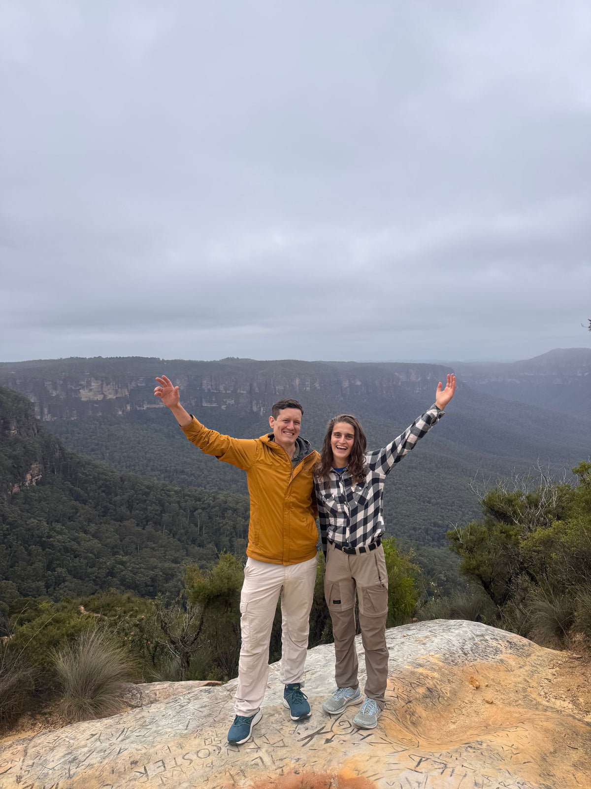 Two people standing on a rocky outcrop with a scenic view of water and trees in the background.